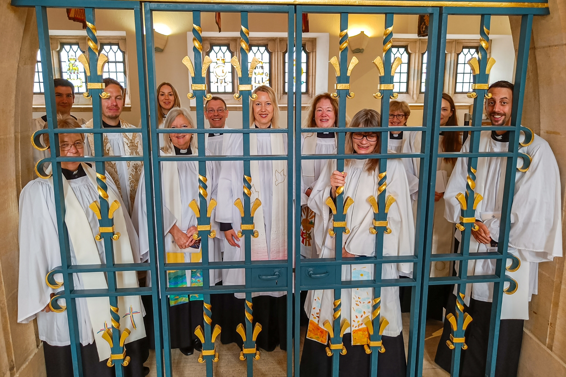 14 new priests behind some ornate bars in Guildford Cathedral in their robes and smiling