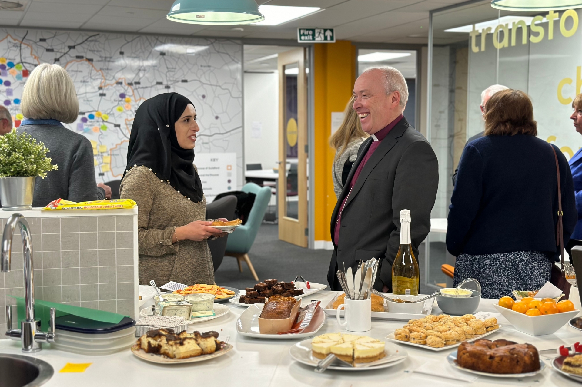 Table full of snack foods and cake. Bishop Paul in conversation with a woman wearing a black head scarf.