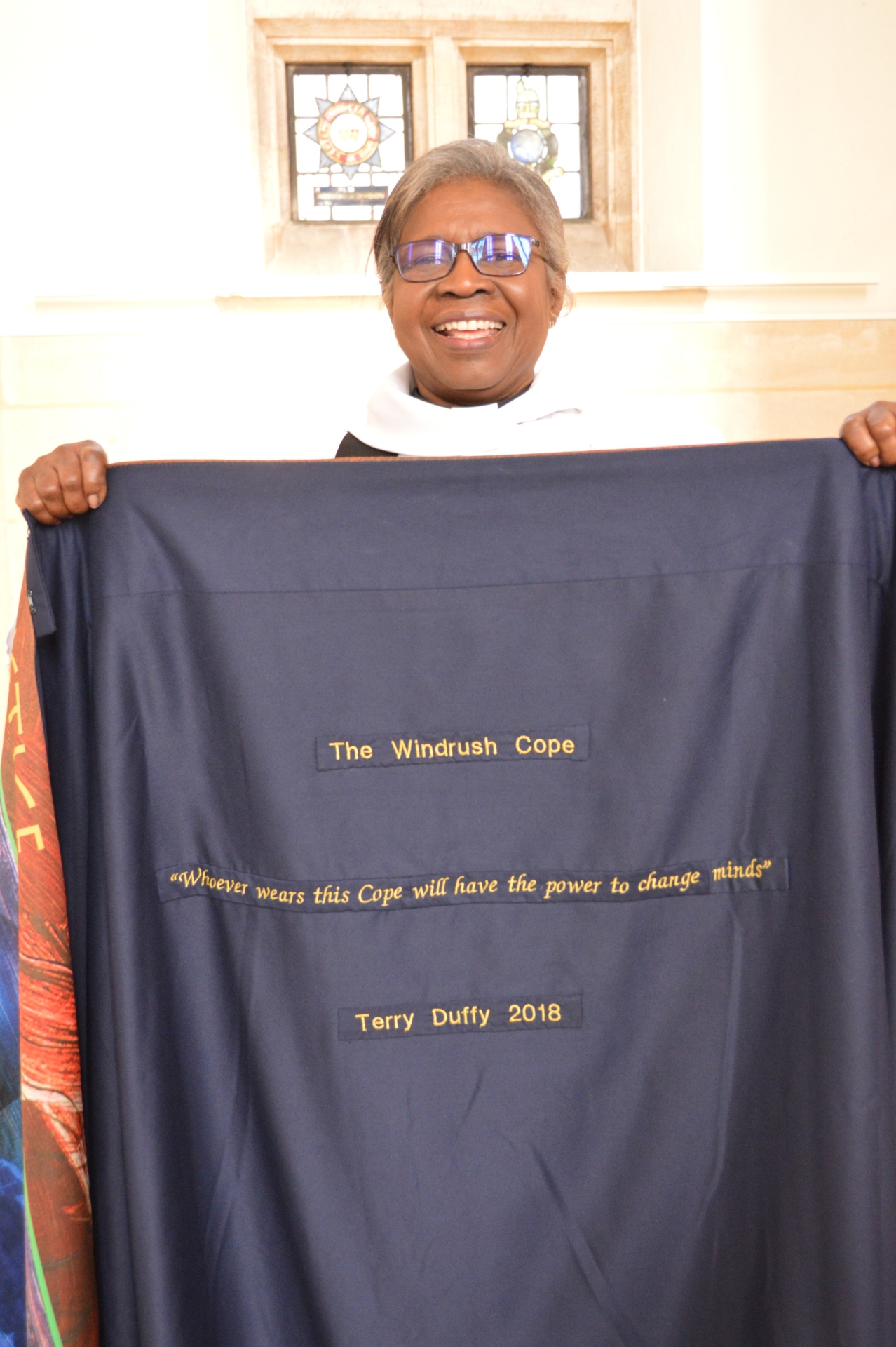 Rev Bev holding up the Windrush Cope which has “Whoever wears this cope will have the power to change minds” embroided on the inside in gold thread on a blue fabric