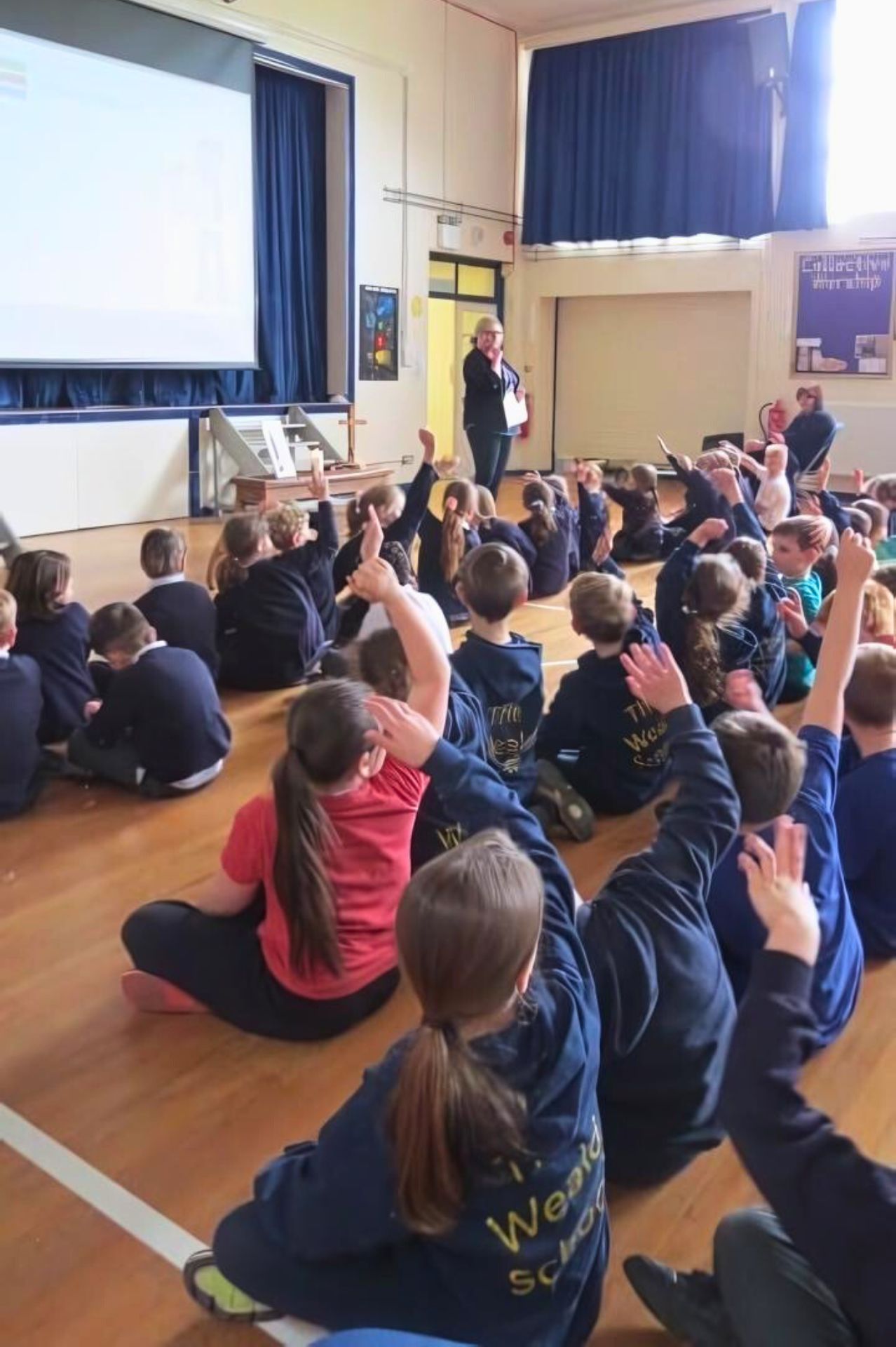 Children sat in an assembly hall with hands raised as a teacher presents at the front.