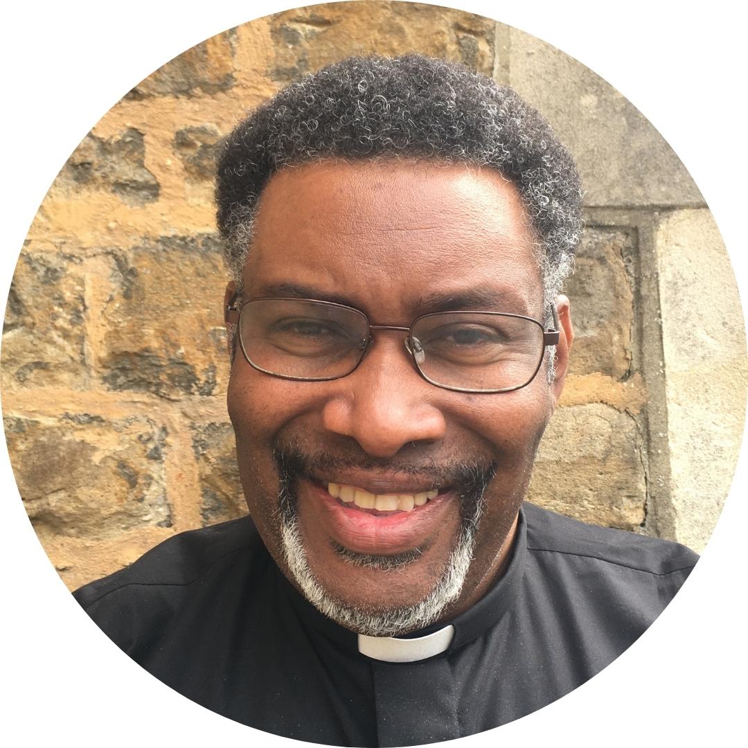 Dean wearing black clerical shirt smiling stood in front of a stone wall