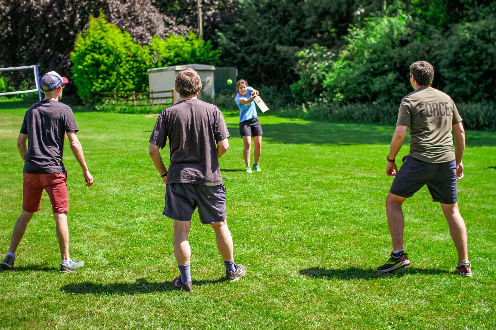 Group of people playing French cricket in a green field surrounded by green trees