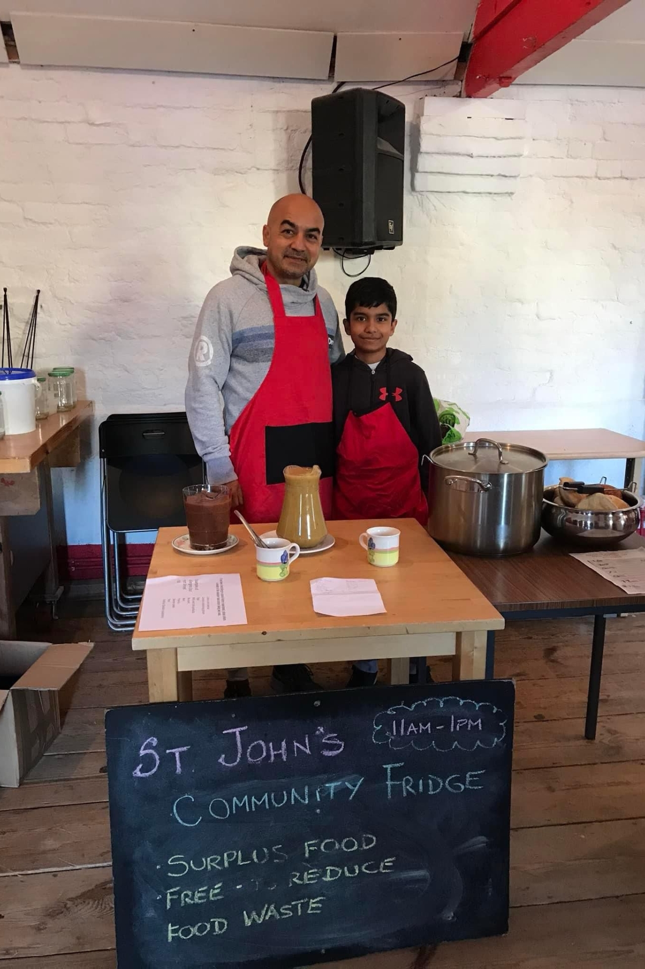 Man and his son wearing red aprons stood behind a chalk written sign reading St John's Community Fridge