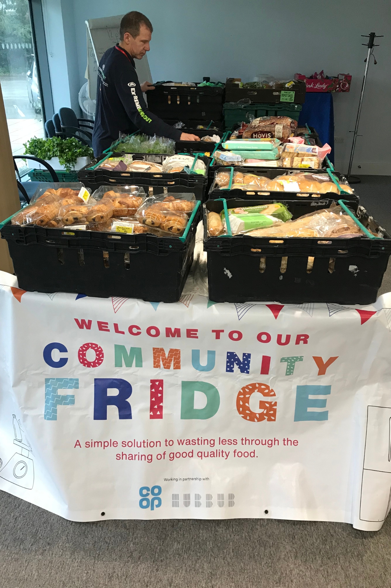 Multiple crates of food on top of a table surrounded by a banner reading Welcome to our Community Fridge