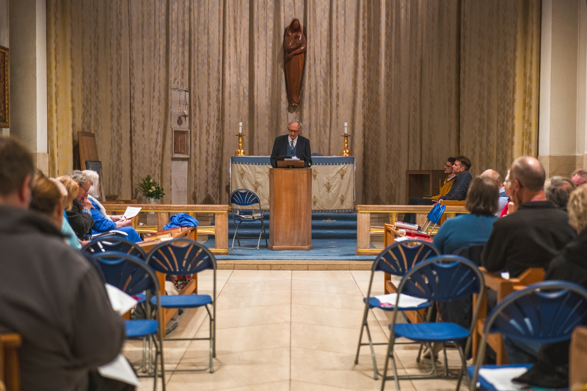 Professor David Ford OBE stood at a lecturn in a chapel giving a lecture whilst people sit looking intently