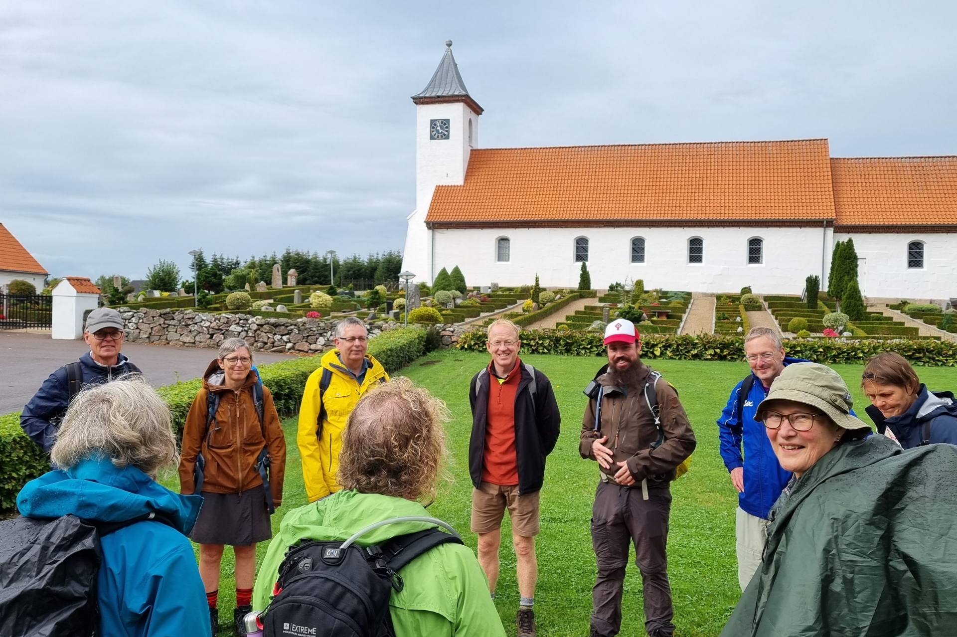Group of people in walking gear gathered on a green outside a church on a cloudy day