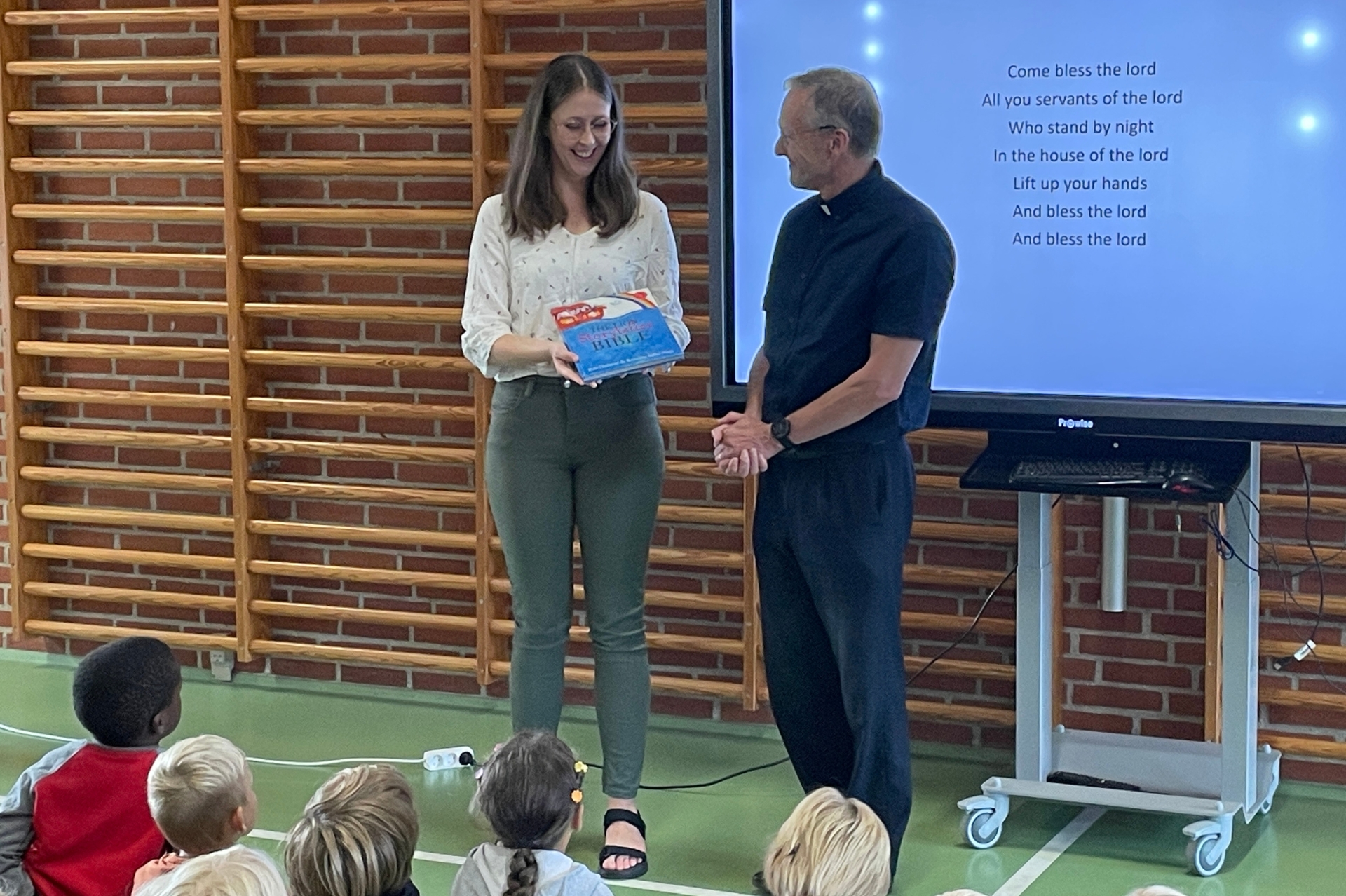 A priest and teacher stood at the front of a school assembly with the teacher presenting a children's book
