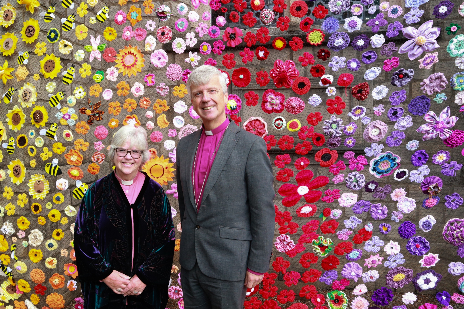 Bishop Andrew and Revd Christine Pattison standing front of the a colourful netting of crafted flowers