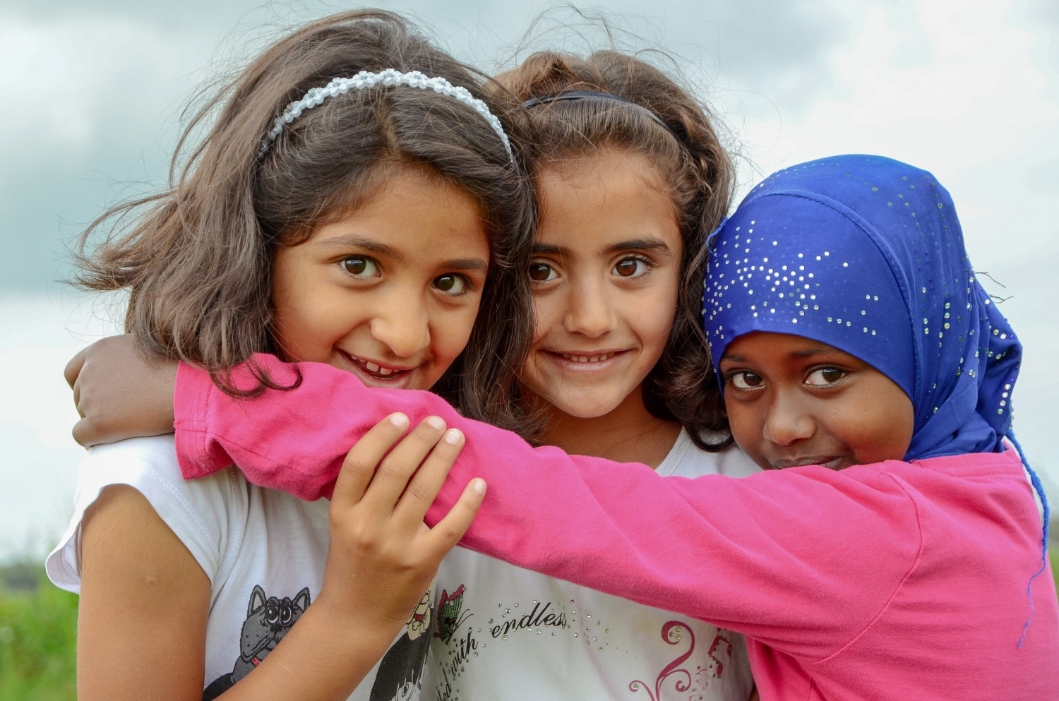 Three young girls, including one wearing a head scarf, hugging and smiling, directly looking at the camera