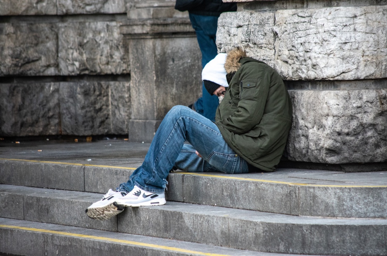 Man wearing a coat, hat, jeans, hoody and white trainers crouched on a concrete step and leaning against a stone wall 