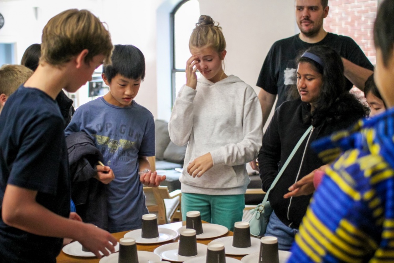 A group of teenagers gathered around a table solving a puzzle involving plates and disposable coffee cups