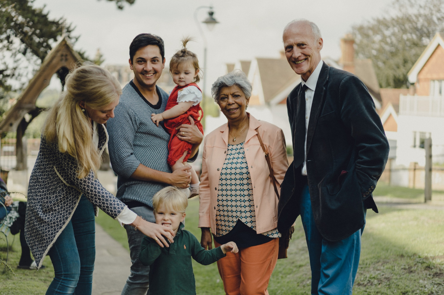 Woman holding young boys hand and man holding young girl in his arms stood next to an other couple in a churchyard all smiling at the camera