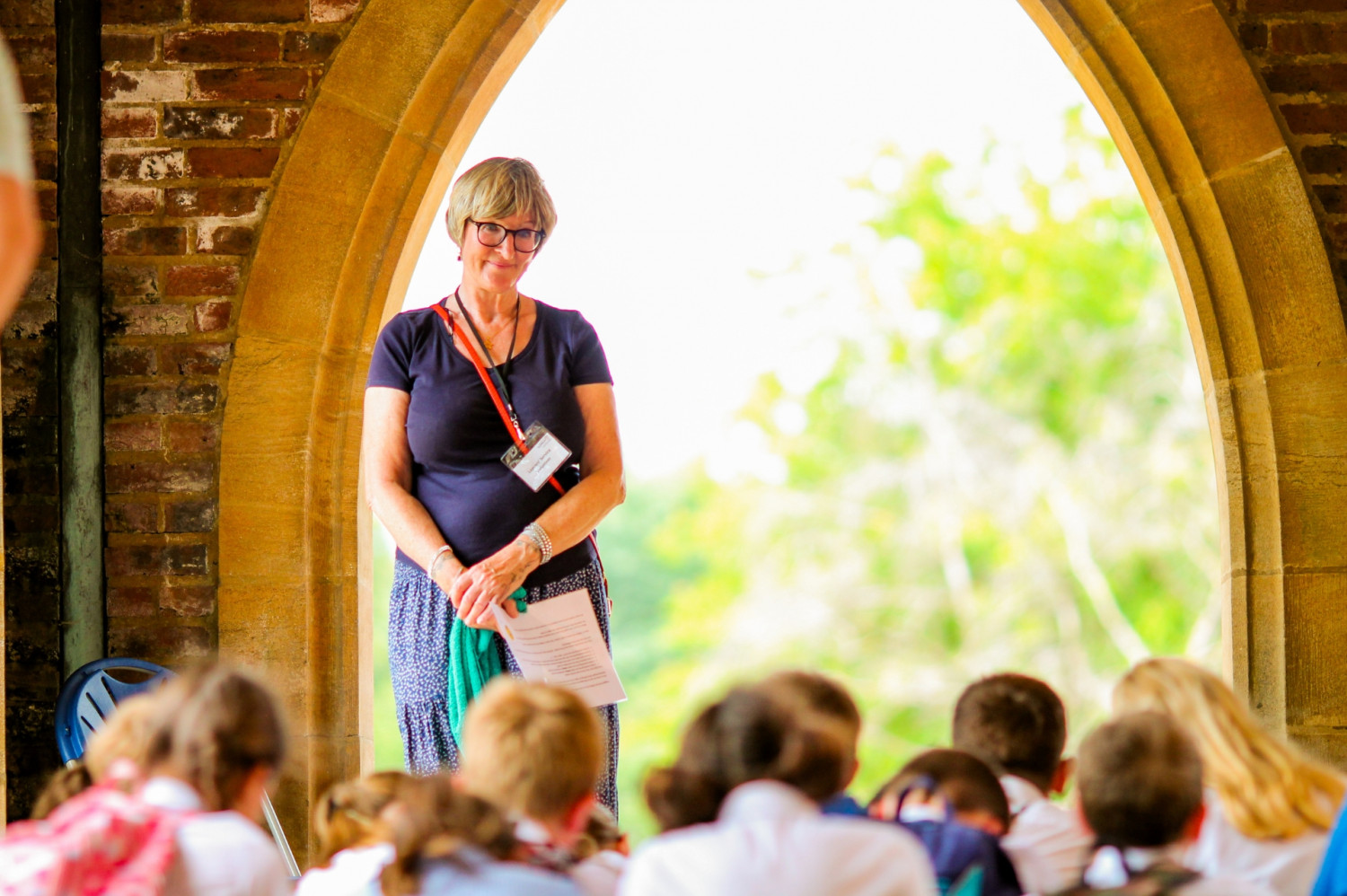 Adult stood up smiling and engaging with school children sat on the floor in an outdoor space