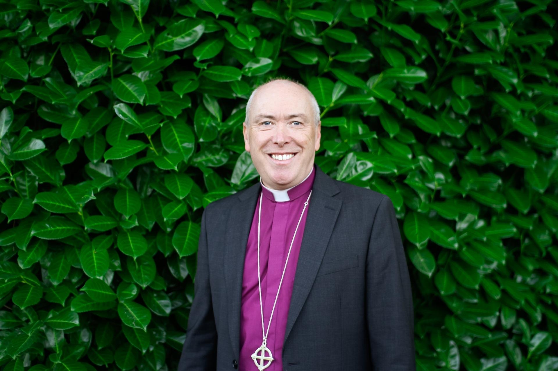 Bishop Paul Davies stood in front of a tall green hedge wearing a purple clerical shirt, silver cross neckless and dark jacket